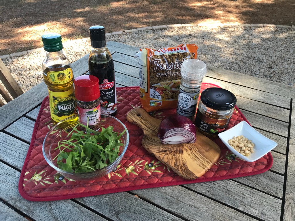 Zutaten auf einem Holztisch unter Pinien: Olivenöl, Crema di Balsamico, Salz, Pfeffermühle, Nudeln, Tomaten, Rucola, Pinienkerne
